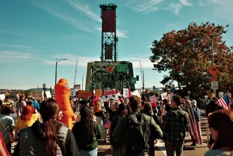 a crowd marching across a portland bridge under its lift tower, one of them in an orange inflatable costume